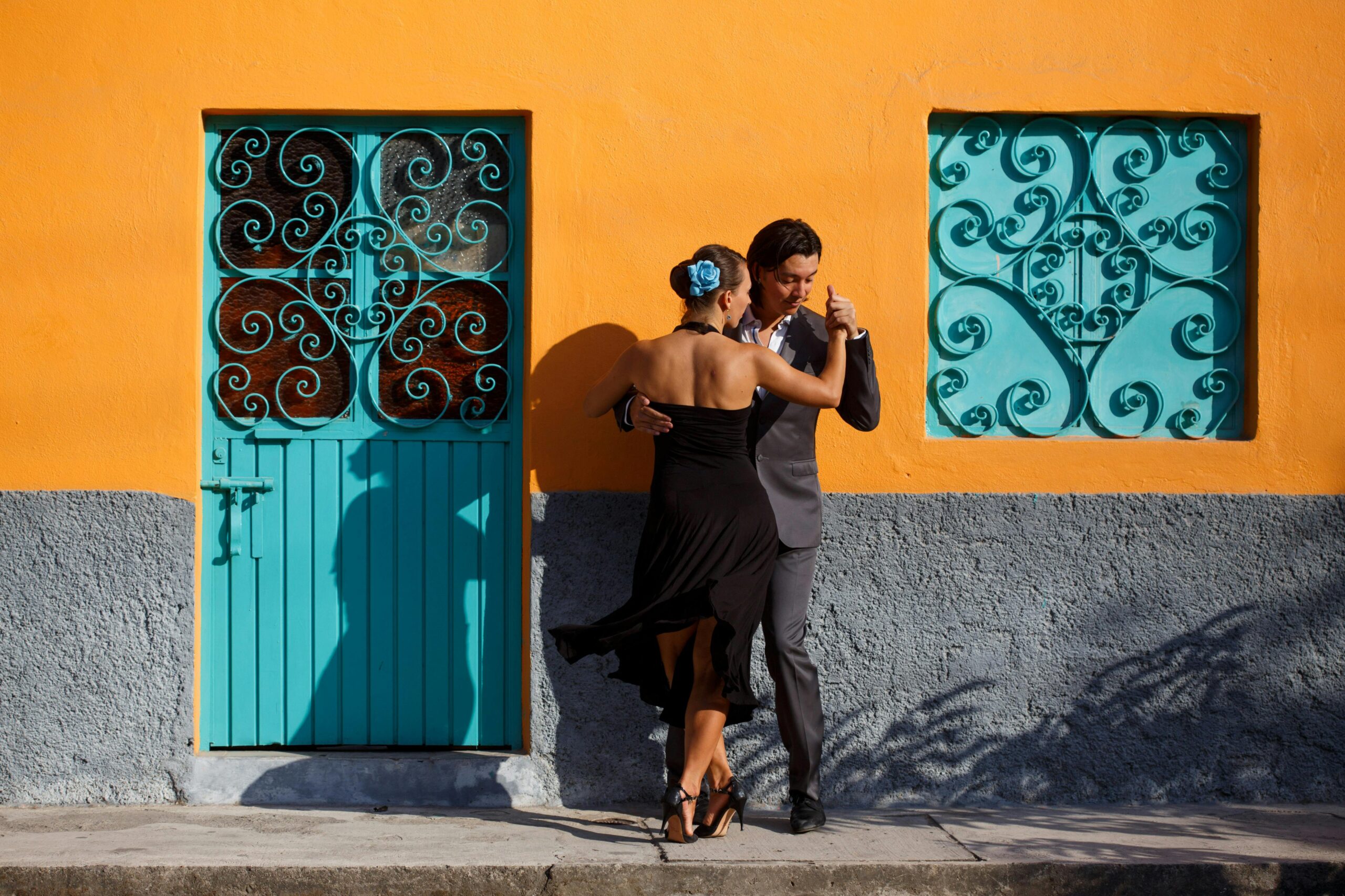 A couple dancing tango in formal attire against a colorful wall with blue accents, showcasing urban cultural expression.